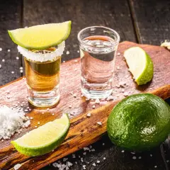 Zwei Tequila-Gläser, Salz und Limettenspalten auf einem Holzbrett. Die Tequila-Reihenfolge: zuerst Salz, dann Tequila, dann Limette.
