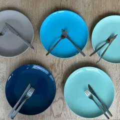 Fünf bunte Teller liegen auf einem Holztisch. Auf jedem Teller liegt besteck auf einer seite des tellers, bereit für ein gemeinsames Essen.