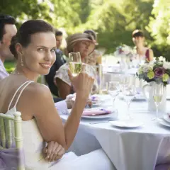 Braut genießt Sekt bei Hochzeitsfeier im Freien. Tischdeko und Gäste im Hintergrund. Guter Knigge bei der Hochzeit ist wichtig.