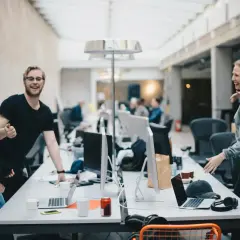 Lachende Kollegen im Büro, die sich austauschen. Der Flurfunk Bedeutung wird hier lebendig, während sie sich über den Schreibtisch beugen.