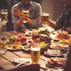 Freunde stoßen mit Bier an, während sie ein Festmahl mit Brathähnchen, Pasta und Salaten genießen. Sie sind zum Essen eingeladen und feiern gemeinsam.