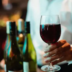 Ein Weinglas mit Rotwein wird gehalten. Im Hintergrund stehen Flaschen. Wo steht das Wasserglas und wo das Weinglas? Hier ist nur das Weinglas zu sehen.