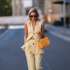 Eine Frau trägt eine elegante gelbe Weste und passende Hose. Sie hält eine auffällige orangefarbene Handtasche.
