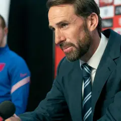 Ein Mann im Anzug mit einem gestreiften Schlips und Kragen spricht in ein Mikrofon. Im Hintergrund ein Spieler im blauen Trainingsanzug.