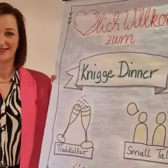 Frau in pinkem Blazer lächelt neben Flipchart mit "Knigge Dinner", Tischkultur und Small Talk.
