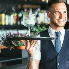 Lächelnder Barkeeper mit Tätowierungen hält ein Tablett mit einem erfrischenden Drink. Er serviert von der rechten Seite.