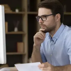 Mann mit Brille am Schreibtisch, der nachdenklich auf den Bildschirm blickt, um alle Mails in Outlook anzuzeigen.