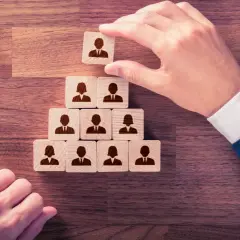 Menschliche Hände ordnen Holzklötze mit Personensymbolen zu einer Pyramide an, die die protokollarische Rangfolge darstellt.