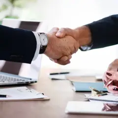 Zwei Geschäftspartner geben sich die **Hand**, um einen Deal abzuschließen. Ein Laptop und Dokumente liegen auf dem Tisch.