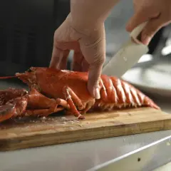 Eine Hand schneidet mit einem Messer einen gekochten Hummer auf einem Holzbrett. So isst man Hummer.