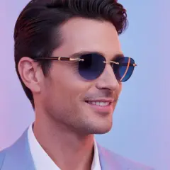 Ein Mann mit stilvoller Sonnenbrille, die den aktuellen Herren-Trend widerspiegelt, lächelt vor einem farbigen Neonlicht.