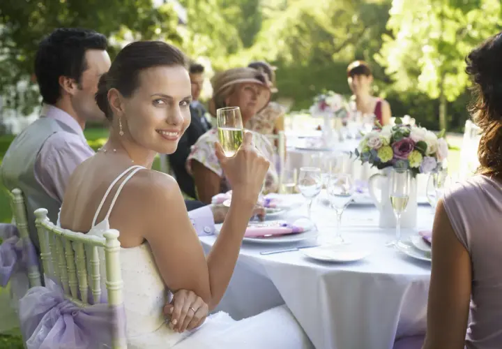 Braut genießt Sekt bei Hochzeitsfeier im Freien. Tischdeko und Gäste im Hintergrund. Guter Knigge bei der Hochzeit ist wichtig.