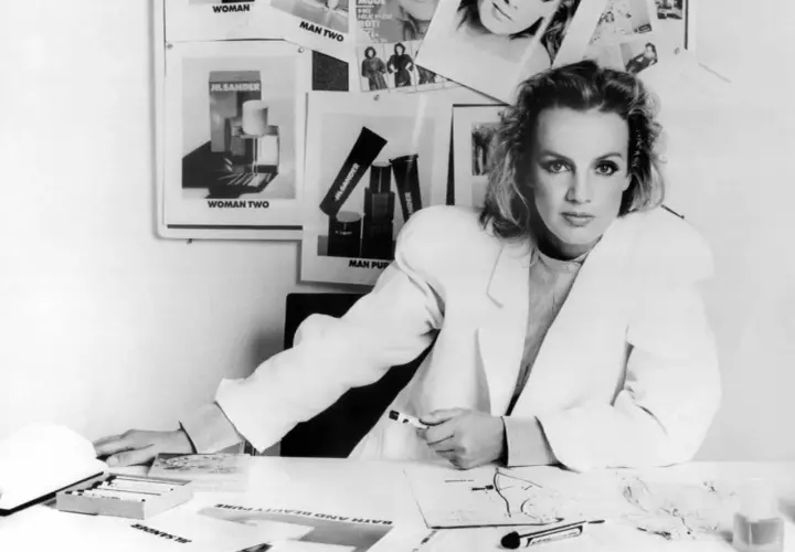 Jil Sander Designerin am Schreibtisch, umgeben von Entwürfen und Produktbildern ihrer Marke.