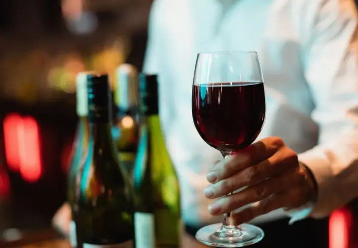 Ein Weinglas mit Rotwein wird gehalten. Im Hintergrund stehen Flaschen. Wo steht das Wasserglas und wo das Weinglas? Hier ist nur das Weinglas zu sehen.