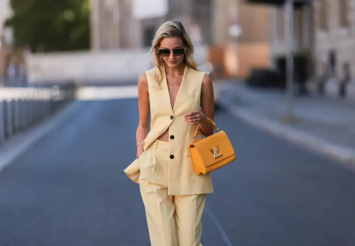 Eine Frau trägt eine elegante gelbe Weste und passende Hose. Sie hält eine auffällige orangefarbene Handtasche.