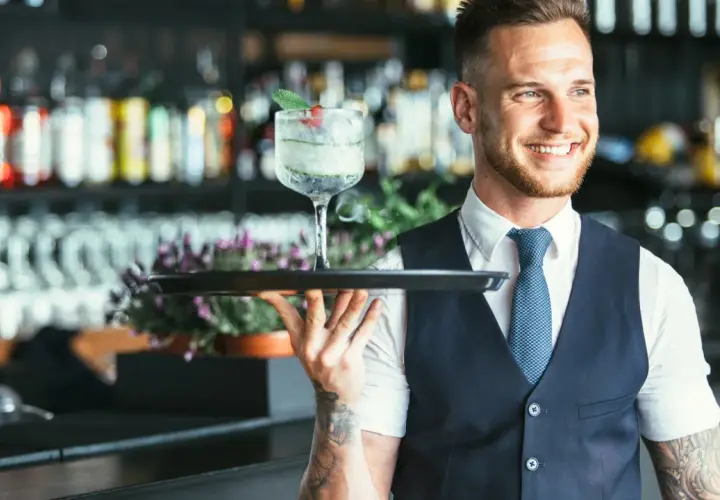 Lächelnder Barkeeper mit Tätowierungen hält ein Tablett mit einem erfrischenden Drink. Er serviert von der rechten Seite.