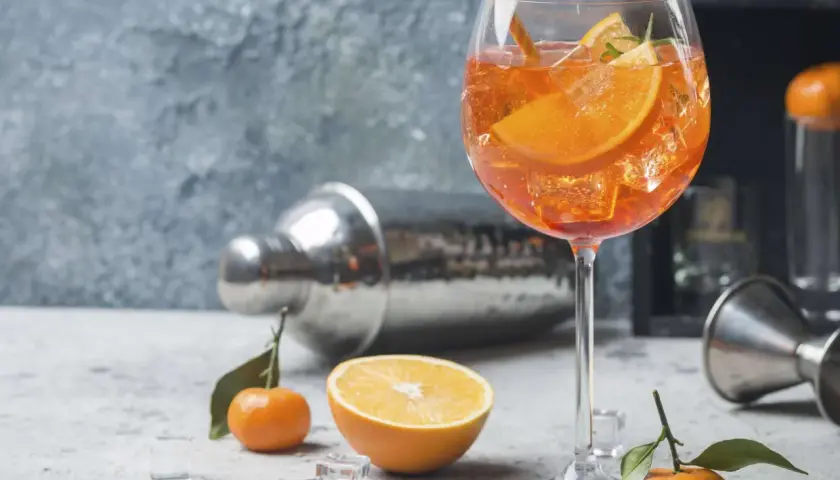 Ein erfrischender Aperitif vor dem Essen: Ein Glas mit einem leuchtend orangen Getränk, Eiswürfeln, Orangenscheiben und einem Strohhalm. Daneben liegen Mandarinen und ein Shaker.