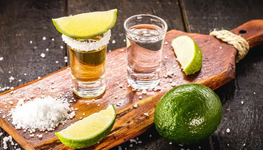 Zwei Tequila-Gläser, Salz und Limettenspalten auf einem Holzbrett. Die Tequila-Reihenfolge: zuerst Salz, dann Tequila, dann Limette.