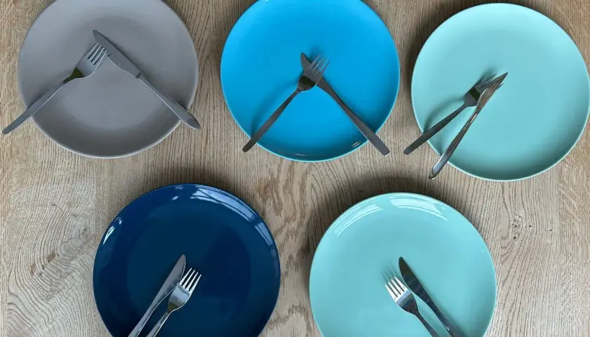 Fünf bunte Teller liegen auf einem Holztisch. Auf jedem Teller liegt besteck auf einer seite des tellers, bereit für ein gemeinsames Essen.