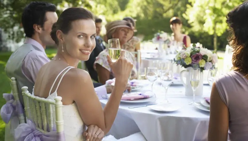 Braut genießt Sekt bei Hochzeitsfeier im Freien. Tischdeko und Gäste im Hintergrund. Guter Knigge bei der Hochzeit ist wichtig.