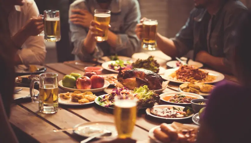 Freunde stoßen mit Bier an, während sie ein Festmahl mit Brathähnchen, Pasta und Salaten genießen. Sie sind zum Essen eingeladen und feiern gemeinsam.