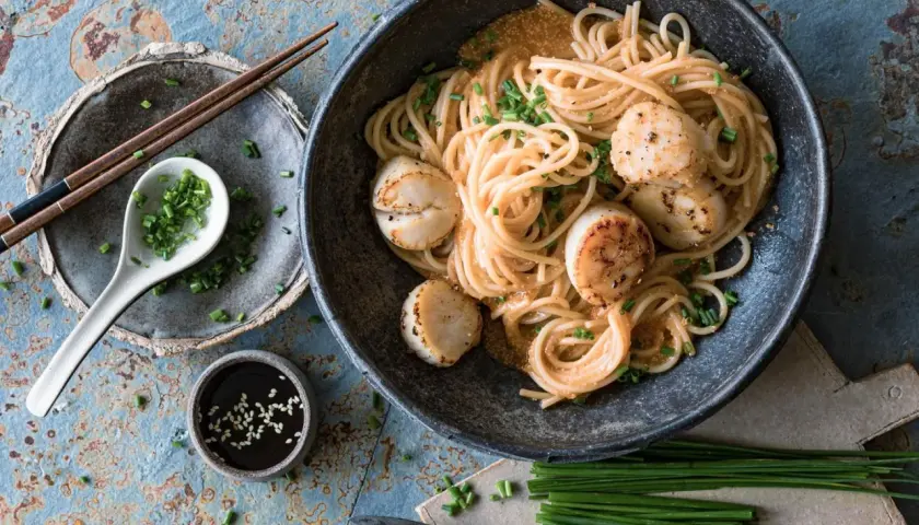 Ein Teller mit Spaghetti und Jakobsmuscheln, garniert mit Schnittlauch. Dazu gibt es Sojasauce und Stäbchen. Ein köstliches Spaghetti-Essen!