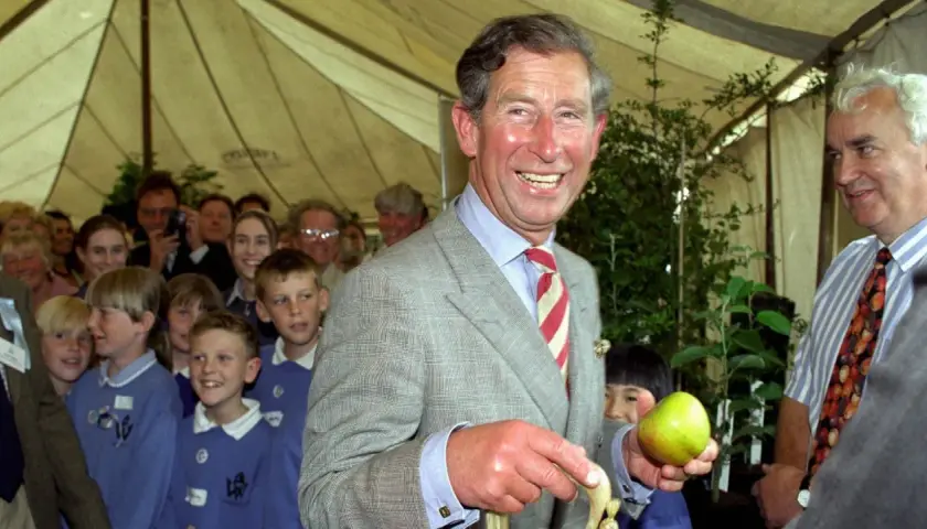 König Charles III. im Prince of Wales Muster-Anzug hält einen Apfel, umgeben von Kindern und Erwachsenen.