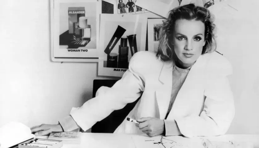 Jil Sander Designerin am Schreibtisch, umgeben von Entwürfen und Produktbildern ihrer Marke.