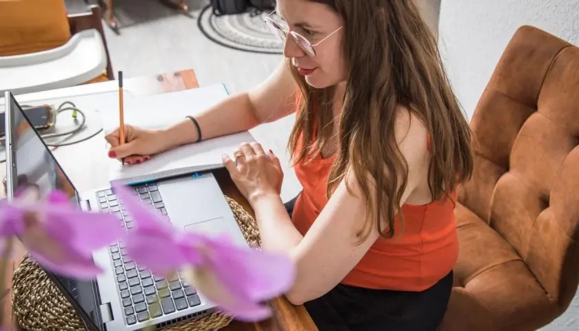 Frau mit Brille schreibt Notizen, während sie am Laptop arbeitet. Sie schafft sich eine gute digitale Ordnung für ihr Homeoffice.