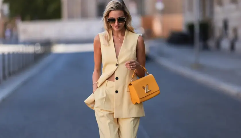Eine Frau trägt eine elegante gelbe Weste und passende Hose. Sie hält eine auffällige orangefarbene Handtasche.