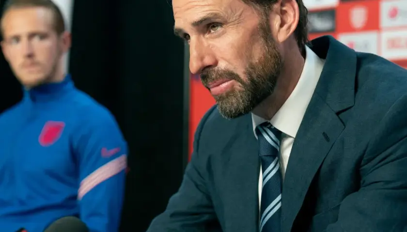 Ein Mann im Anzug mit einem gestreiften Schlips und Kragen spricht in ein Mikrofon. Im Hintergrund ein Spieler im blauen Trainingsanzug.