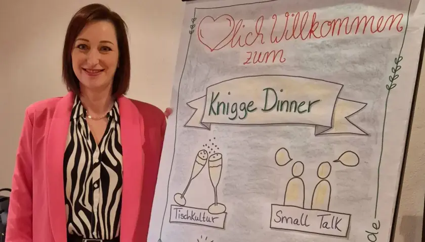 Frau in pinkem Blazer lächelt neben Flipchart mit "Knigge Dinner", Tischkultur und Small Talk.