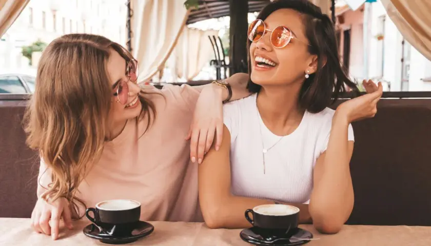 Zwei Freundinnen lachen bei Kaffee. Sie zeigen, wie gut man auf Komplimente reagiert: mit Freude und Selbstbewusstsein.