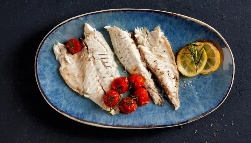 Drei zarte Fischfilets liegen auf einem blauen Teller, garniert mit Kirschtomaten und Zitronenscheiben. Perfekt zum Fisch filetieren am Tisch.