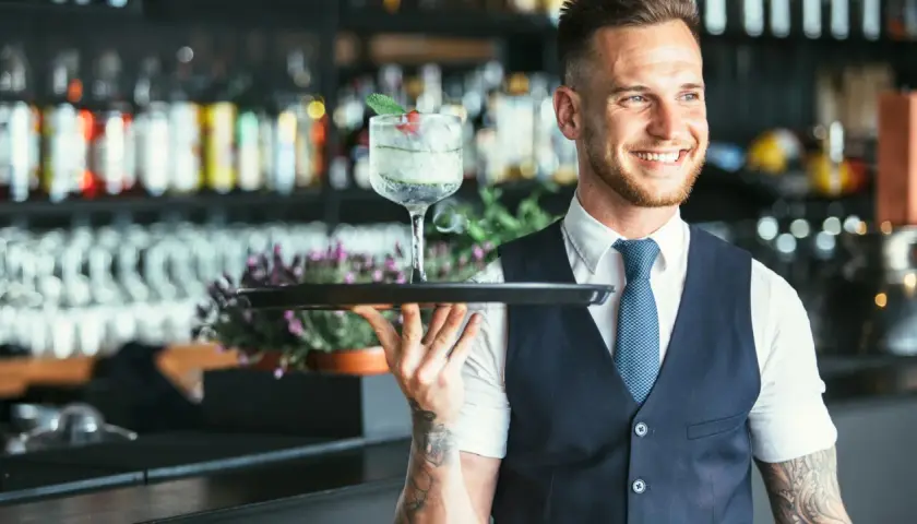 Lächelnder Barkeeper mit Tätowierungen hält ein Tablett mit einem erfrischenden Drink. Er serviert von der rechten Seite.