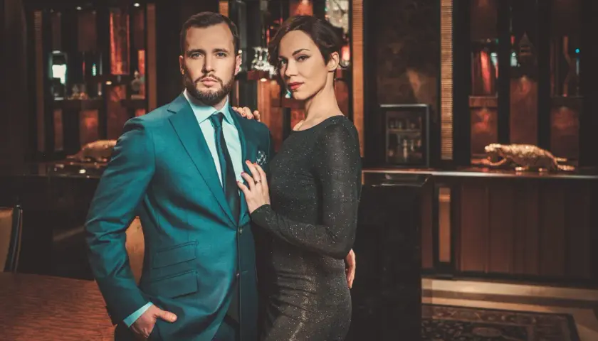 Ein eleganter englischer Gentleman im Anzug posiert mit einer Frau in einem glitzernden Kleid in einem luxuriösen Interieur.