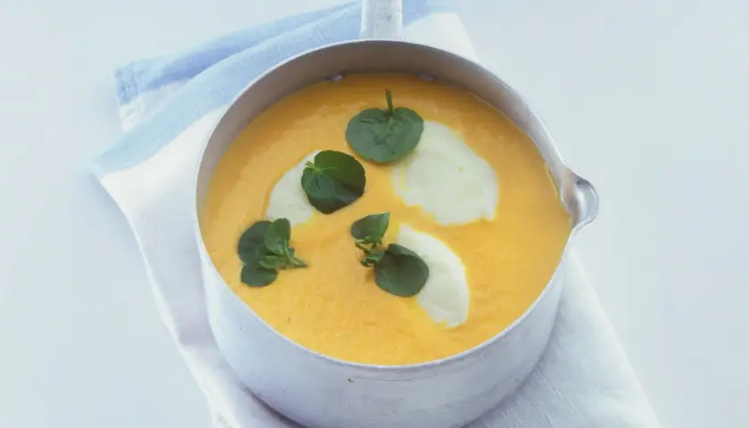 Eine cremige, orangefarbene Suppe mit Klecksen von weißer Creme und grünen Blättern, bereit zum Suppe essen.