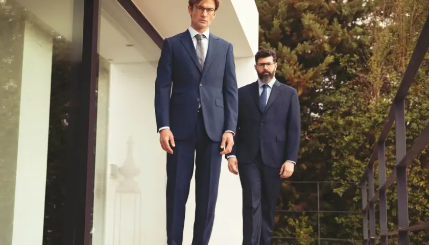 Zwei Männer in eleganten Anzügen stehen auf einer Holzterrasse. Sie zeigen, welche Schuhe zum Anzug passen: klassische Lederschuhe.