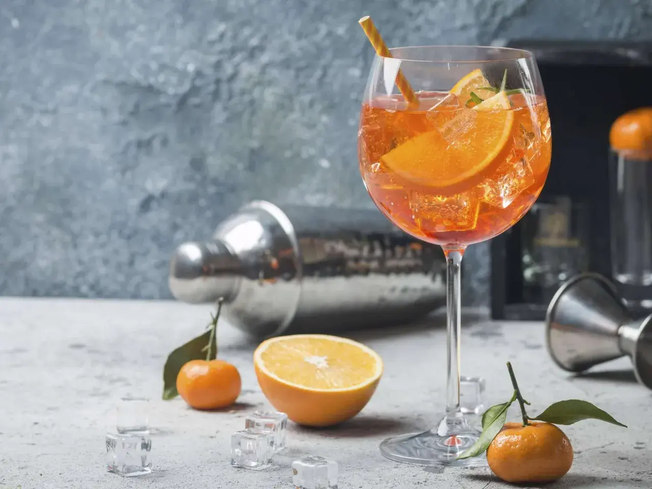 Ein erfrischender Aperitif vor dem Essen: Ein Glas mit einem leuchtend orangen Getränk, Eiswürfeln, Orangenscheiben und einem Strohhalm. Daneben liegen Mandarinen und ein Shaker.