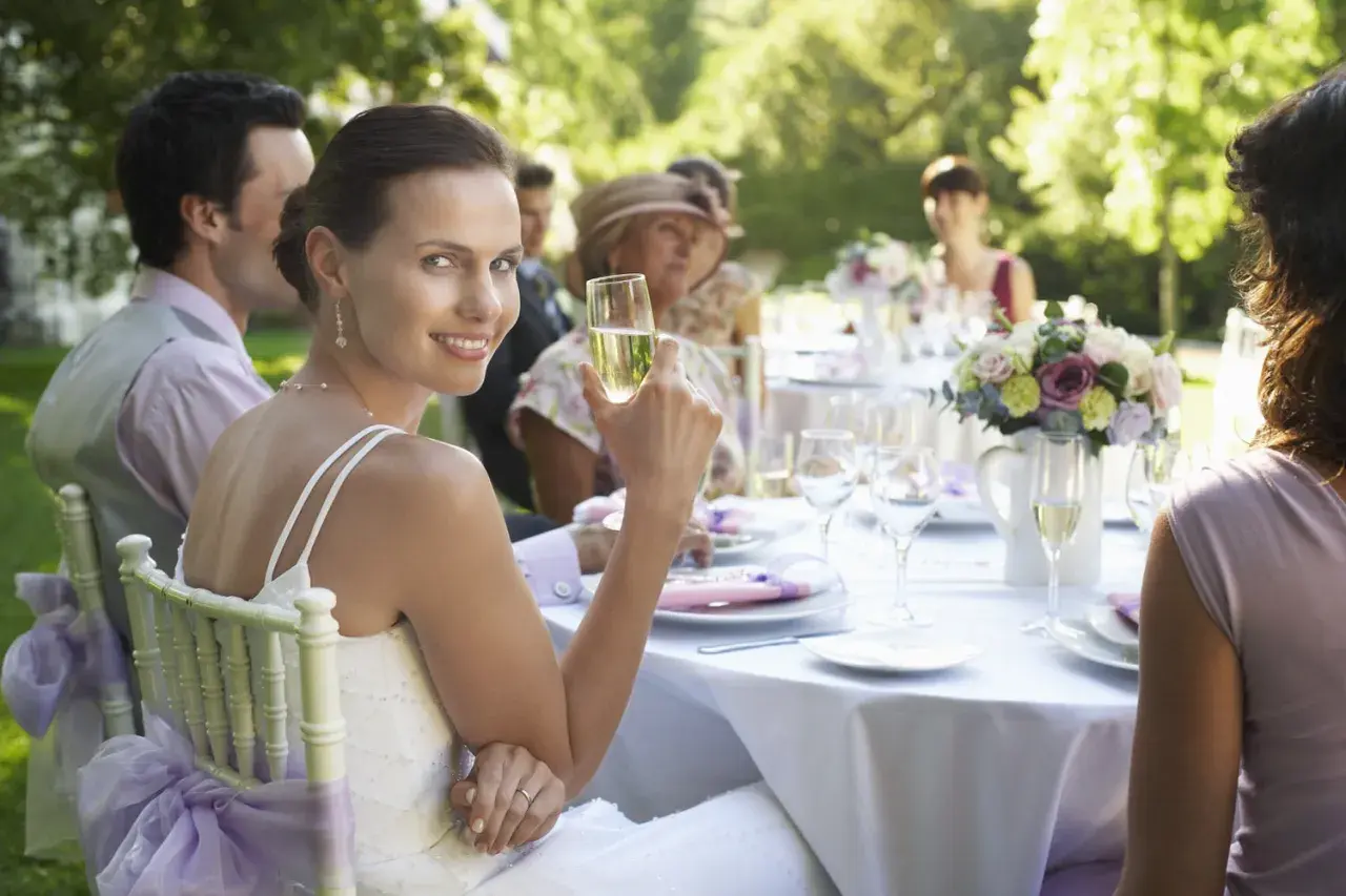 Braut genießt Sekt bei Hochzeitsfeier im Freien. Tischdeko und Gäste im Hintergrund. Guter Knigge bei der Hochzeit ist wichtig.
