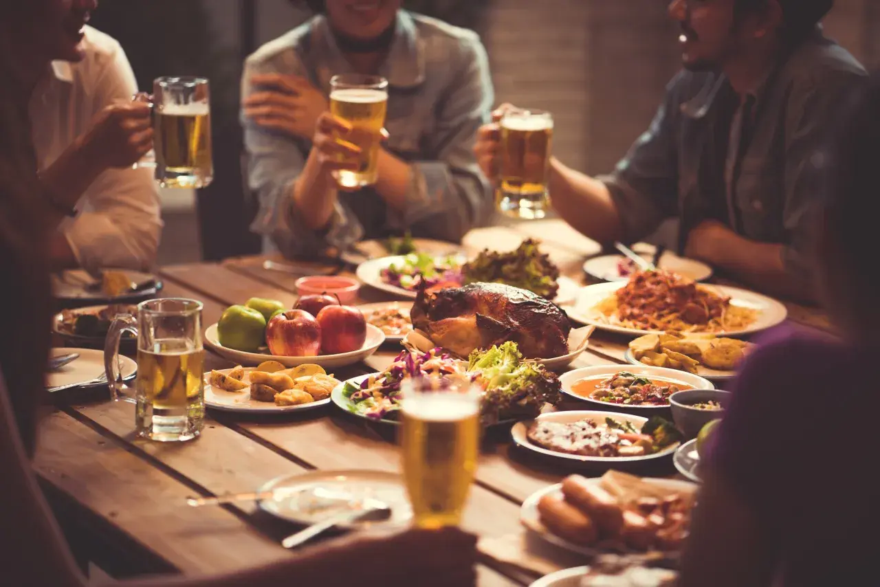 Freunde stoßen mit Bier an, während sie ein Festmahl mit Brathähnchen, Pasta und Salaten genießen. Sie sind zum Essen eingeladen und feiern gemeinsam.