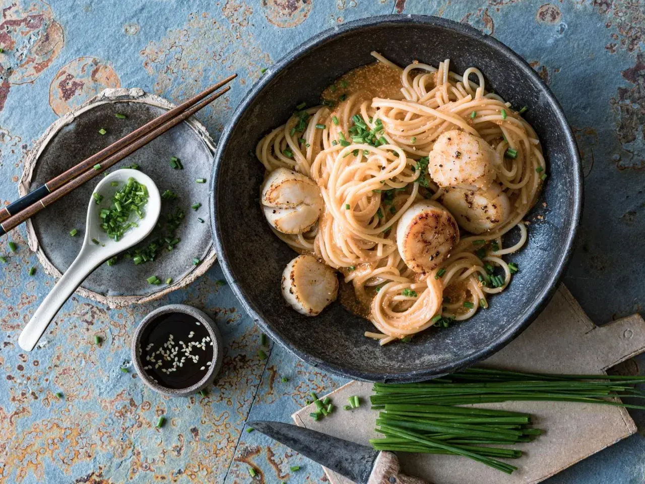 Ein Teller mit Spaghetti und Jakobsmuscheln, garniert mit Schnittlauch. Dazu gibt es Sojasauce und Stäbchen. Ein köstliches Spaghetti-Essen!