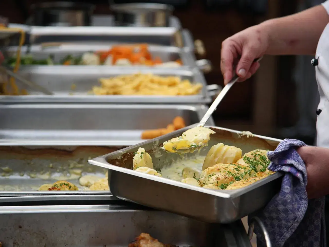 Eine Hand schöpft Kartoffeln aus einer Chafing Dish. Da fragt man sich, warum sagt man Mahlzeit? Leckeres Essen für alle!