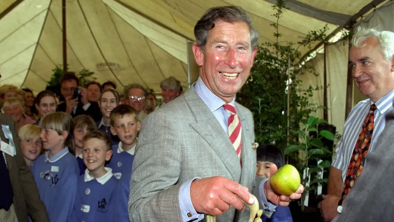 König Charles III. im Prince of Wales Muster-Anzug hält einen Apfel, umgeben von Kindern und Erwachsenen.