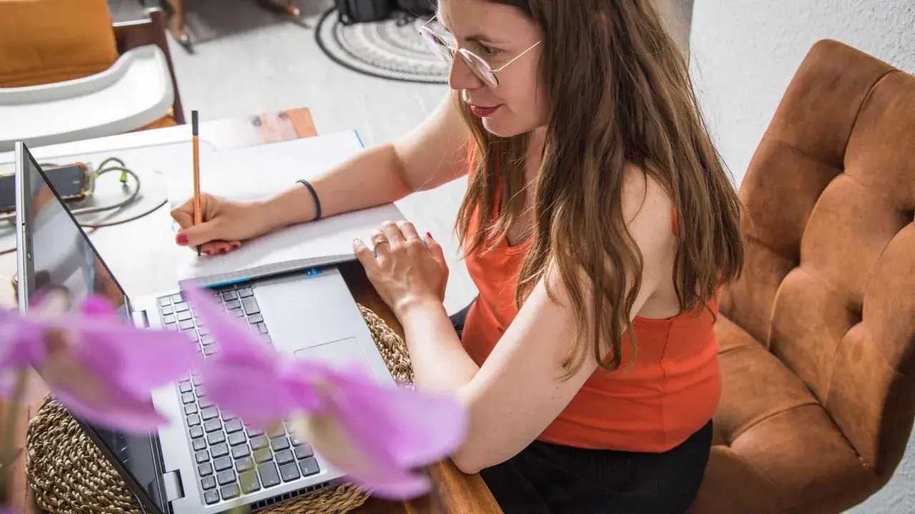 Frau mit Brille schreibt Notizen, während sie am Laptop arbeitet. Sie schafft sich eine gute digitale Ordnung für ihr Homeoffice.