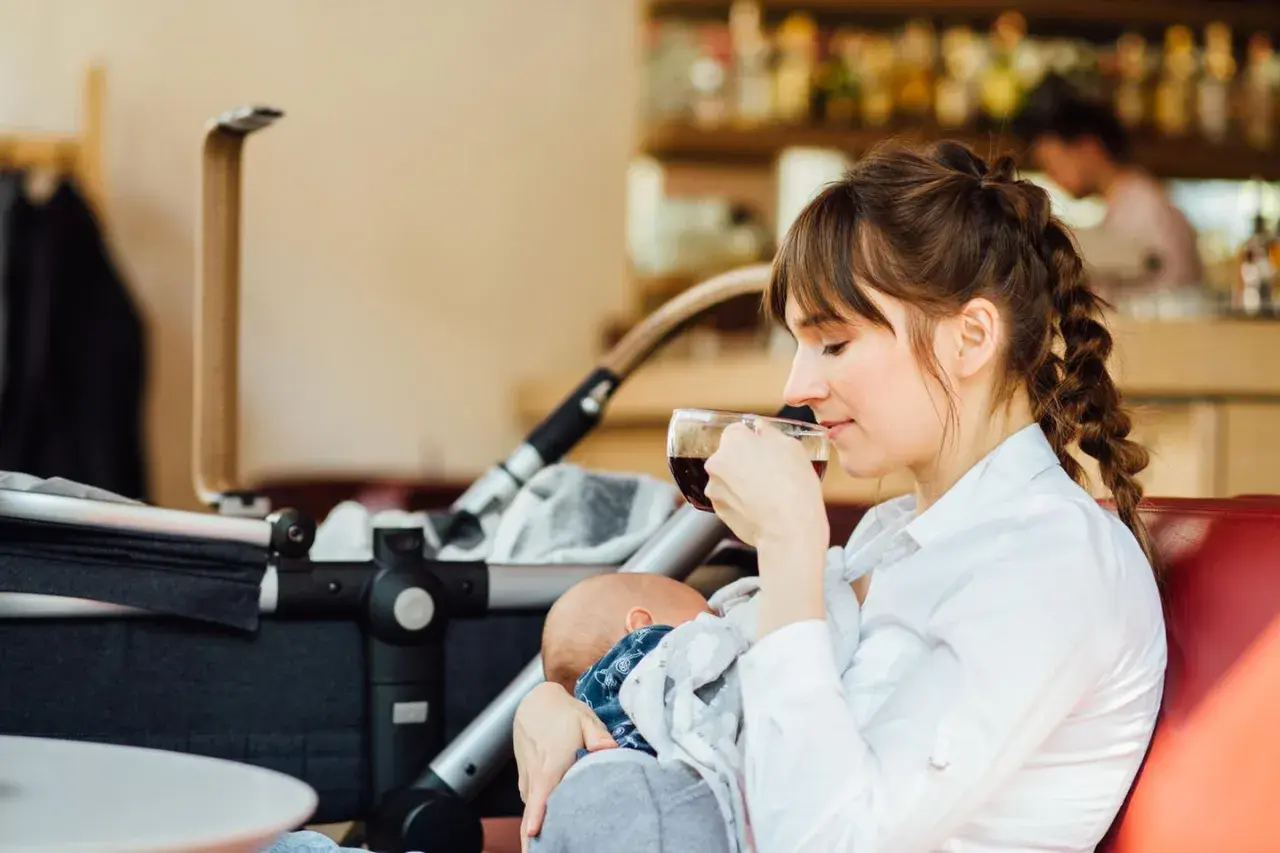Eine junge Mutter genießt eine Tasse Kaffee, während sie ihr Baby stillt. Der Kinderwagen steht daneben. Ein Moment der Ruhe beim Stillen im Restaurant.