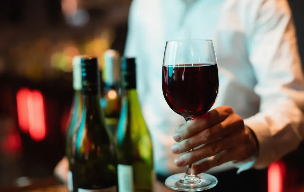 Ein Weinglas mit Rotwein wird gehalten. Im Hintergrund stehen Flaschen. Wo steht das Wasserglas und wo das Weinglas? Hier ist nur das Weinglas zu sehen.