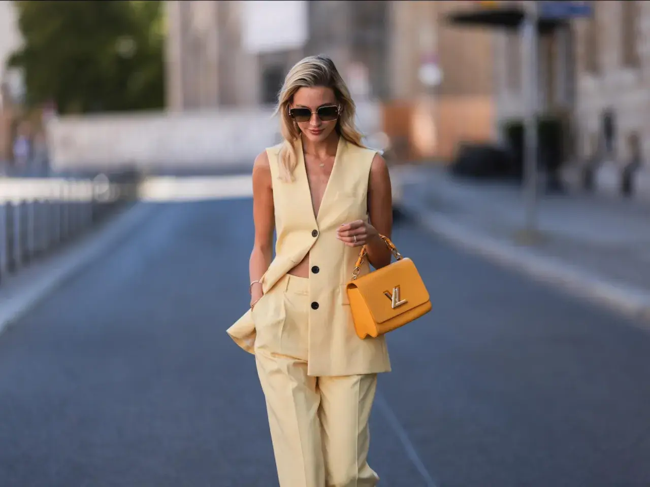 Eine Frau trägt eine elegante gelbe Weste und passende Hose. Sie hält eine auffällige orangefarbene Handtasche.