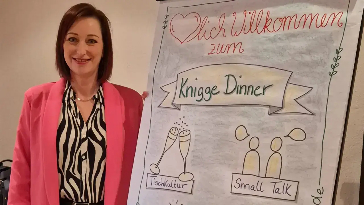 Frau in pinkem Blazer lächelt neben Flipchart mit "Knigge Dinner", Tischkultur und Small Talk.