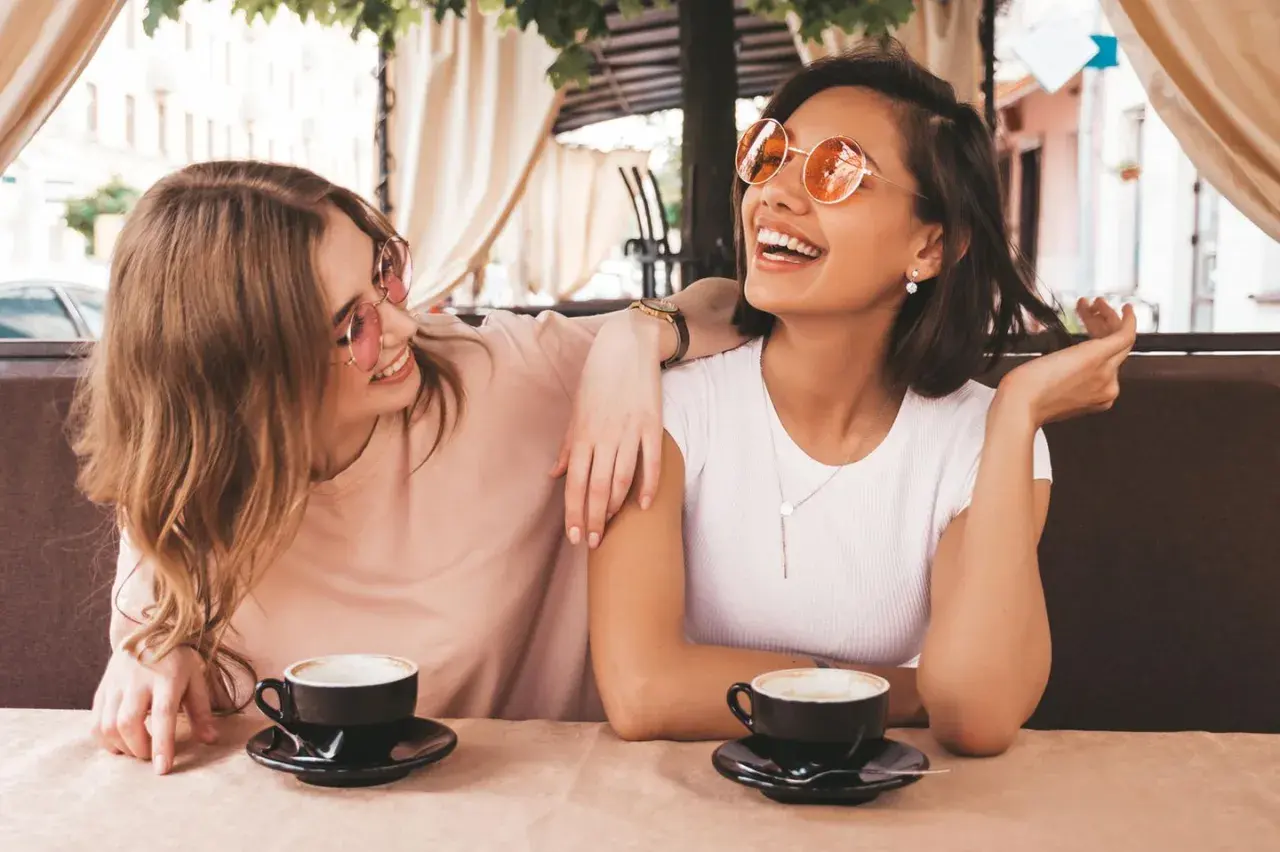Zwei Freundinnen lachen bei Kaffee. Sie zeigen, wie gut man auf Komplimente reagiert: mit Freude und Selbstbewusstsein.